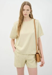 Lichtbeige, losvallend T-shirt met korte mouwen gecombineerd met gestructureerde lichte shorts. Model houdt een bruine schoudertas vast. Eenvoudig ontwerp en zachte stof.