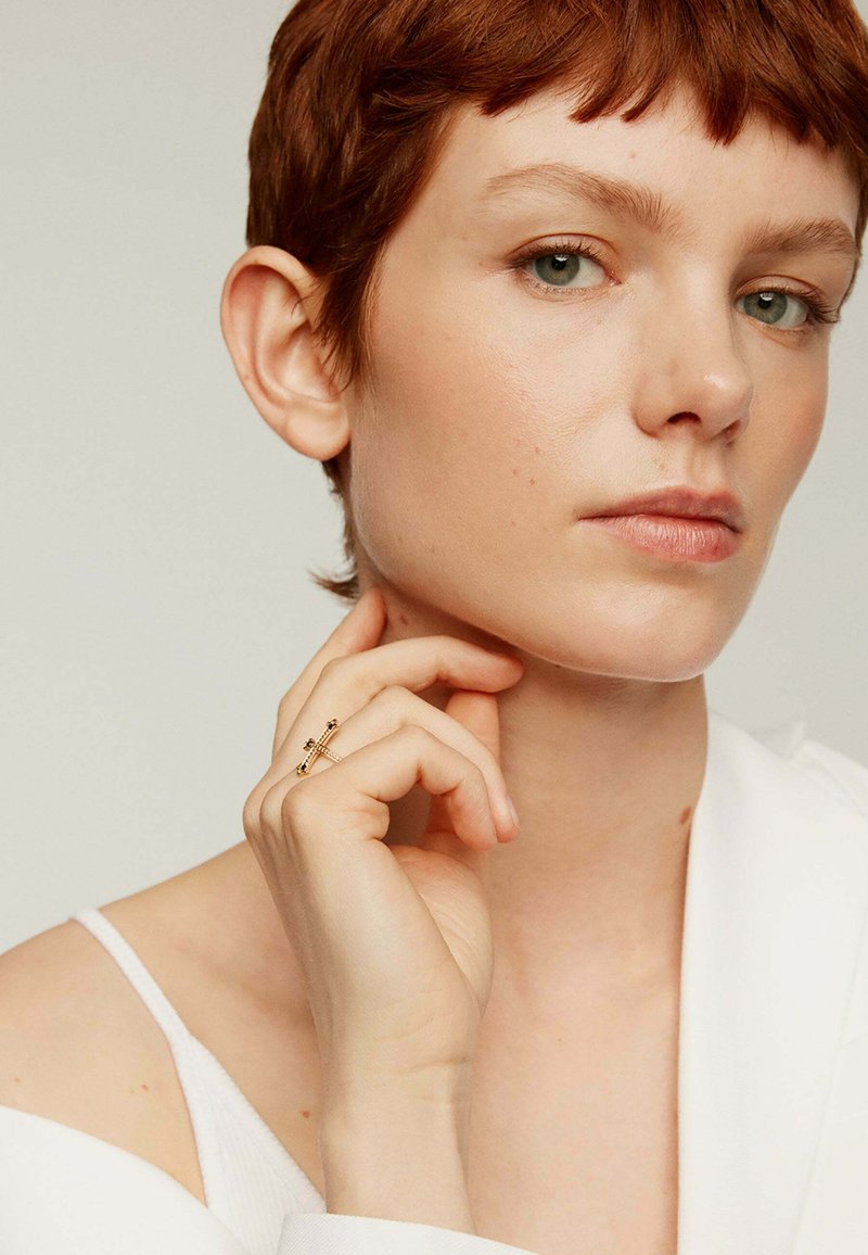 Anillo de oro con acentos negros y piedras transparentes, llevado en una mano, que muestra un diseño delicado y el tono de piel. El modelo tiene el cabello rojo corto y una blusa blanca.