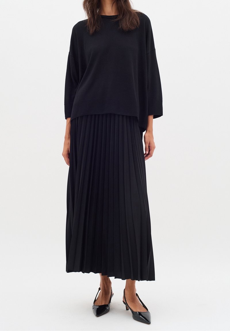 Femme portant un pull ample noir, une longue jupe plissée noire et des escarpins slingback en cuir verni noir, sur un fond blanc uni.