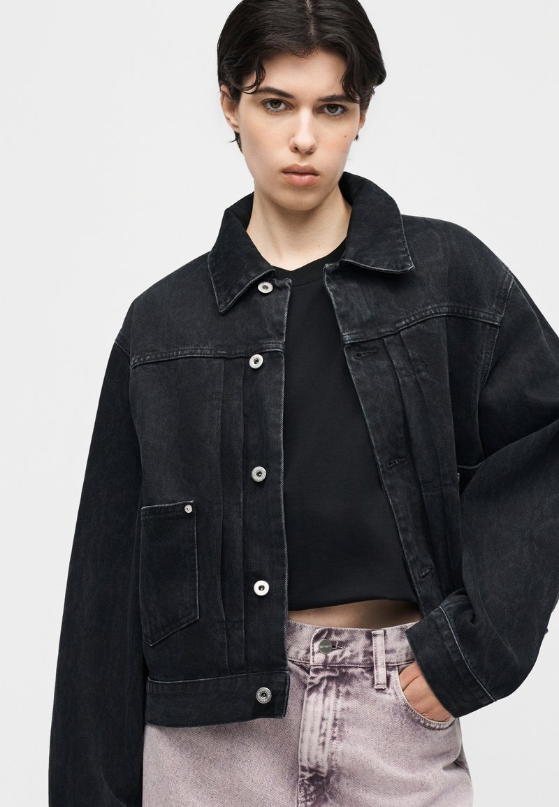 Giovane che indossa una giacca di jeans nera oversize sopra un top corto nero e jeans a vita alta viola chiaro, in piedi con una mano in tasca.