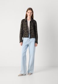 Zwarte zip-up jas met een groene bloemenpatroon, gecombineerd met lichtblauwe wijde jeans en beige spitse schoenen op een effen achtergrond.