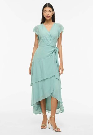 Kvinde iført en lyseblå slå-om-kjole med flagrende ærmer og lagdelt asymmetrisk kant, kombineret med guld høje hæl sandaler.