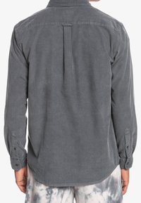 Szara koszula z długim rękawem, z kordurojowego materiału, z plisą z tyłu, mankietami zapinanymi na guziki i zaokrąglonym dołem. Tkanina ma miękką, teksturowaną powierzchnię.