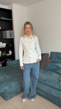 Femme debout dans le salon portant une veste blanche boutonnée, un jean large bleu, des baskets blanches, à côté d'un canapé en velours côtelé bleu canard avec des coussins.