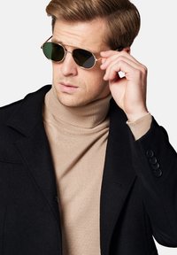 Okrągłe, złote okulary przeciwsłoneczne z zielonymi soczewkami. Noszone przez osobę w czarnym płaszczu na beżowym golfie. Gładkie faktury i dopasowany krój.