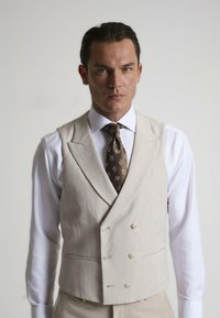 Chaleco de lino beige con un diseño de doble botonadura, que cuenta con dos bolsillos frontales y botones redondos a juego, combinado con una camisa blanca y una corbata estampada.