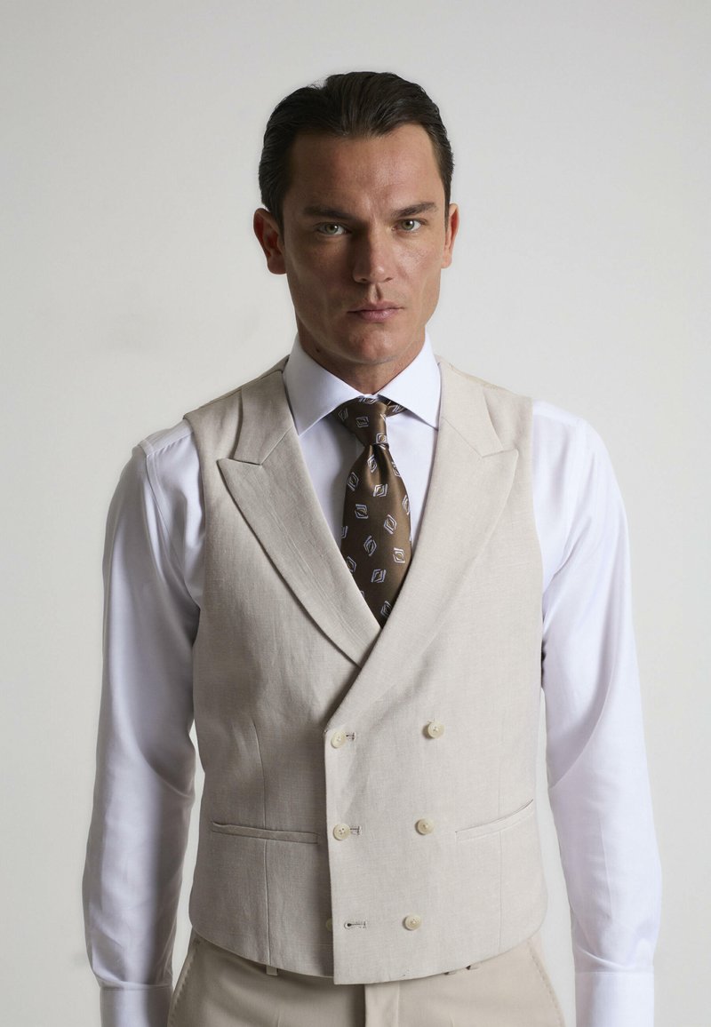 Chaleco de lino beige con un diseño de doble botonadura, que cuenta con dos bolsillos frontales y botones redondos a juego, combinado con una camisa blanca y una corbata estampada.