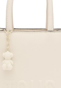 Bolso de mano texturizado blanco con dos asas cortas, que presenta un pequeño charm de oso blanco sujeto por una cadena. Letras visibles en la parte frontal.