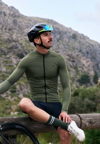 Jersey de ciclismo ajustado de manga larga en color verde oliva con cremallera frontal, combinado con pantalones cortos negros y calcetines a rayas verdes, acompañado de un casco negro.