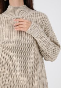 Pull beige en maille avec un col montant côtelé, coupe ample et motif texturé ; manches longues avec poignets côtelés.