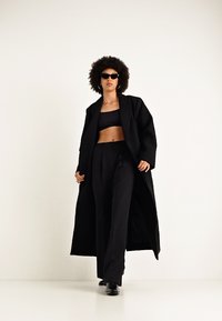 Sort skræddersyet frakke, matchende bredbenede bukser og en cropped top; modellen bærer solbriller. Glatte teksturer, minimalistisk design og rene linjer.