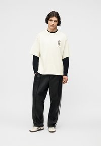 Νέος άνθρωπος με μεσαίου μήκους σκούρα μαλλιά, φορώντας λευκό κοντομάνικο πουκάμισο πάνω από μαύρο μακρυμάνικο τοπ, μαύρο παντελόνι και λευκά αθλητικά παπούτσια.