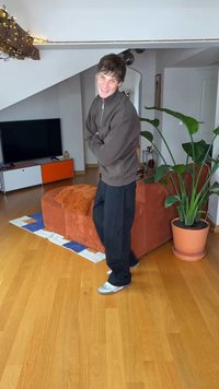 Ruda užtraukiama striukė, juodos kelnės, pilki batai, stovintis kambaryje su medinėmis grindimis, oranžiniu sofa ir dideliu žaliu augalu vazonėlyje.