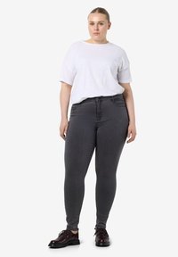 Mujer con el cabello rubio recogido, vistiendo una camisa blanca holgada de manga corta, pantalones ajustados oscuros y zapatos marrones oscuros, de pie mirando hacia adelante.