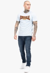 Šviesiai mėlyna marškinėliai su ryškiu raudonu ir geltonu „LONSDALE LONDON“ logotipu. Priglundantį dizainą derinkite su tamsiai mėlynomis džinsomis ir baltais sportbačiais.
