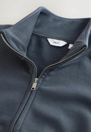 Dunkelgrau zip-up Jacke mit hohem Kragen, glattem Stoff und metallischem Reißverschluss, mit einem sichtbaren Etikett mit der Aufschrift "NEXT".