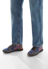 Blaue und graue Sneaker mit roten Akzenten, die mit blauen Jeans auf einer weißen Oberfläche stehen und Schatten werfen.