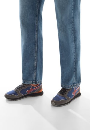 Sneaker blu e grigi con dettagli rossi indossati con jeans blu, posizionati su una superficie bianca e che proiettano ombre.