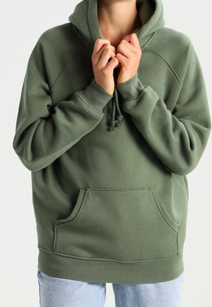 Personne portant un sweat à capuche vert olive avec poche avant et cordons, tenant la capuche près du cou, associée à un jean bleu clair.