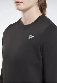 Femme portant un sweat-shirt noir à col rond Reebok, montrant le haut du torse et le cou dans une pose neutre sur fond uni.