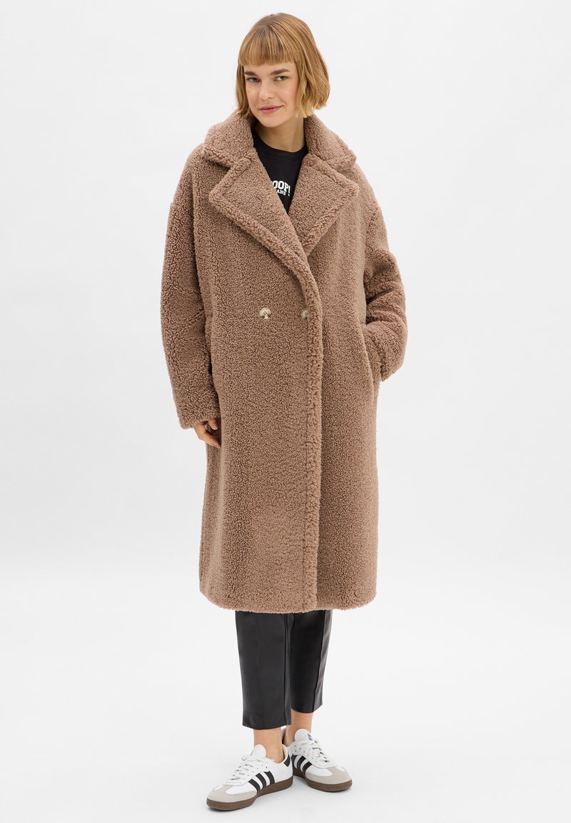 Brauner Lammfell-Mantel mit oversized Passform, Reverskragen, doppelt-knöpfender Verschluss und seitlichen Taschen; kombiniert mit schwarzen Hosen und Sneakers.