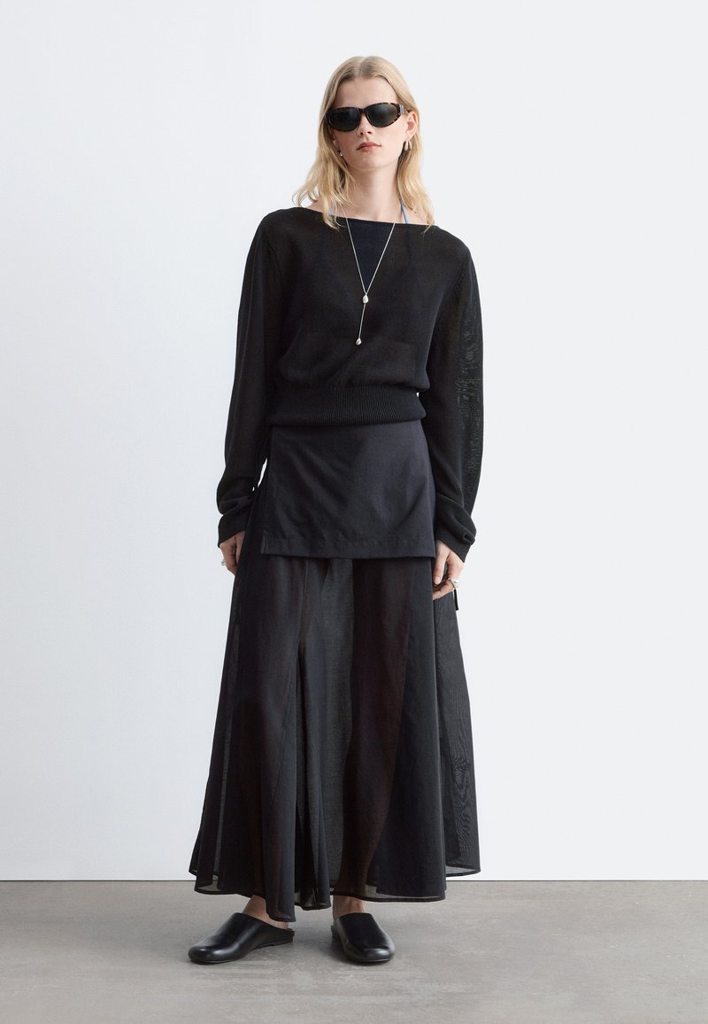 Femme portant un haut noir à manches longues, une jupe noire superposée, des chaussures noires sans lacets, des lunettes de soleil et un long collier pendentif, debout devant un fond uni.