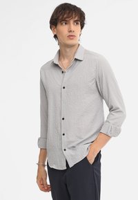 Camisa de manga larga gris claro con tejido texturizado, cuello con botones, botones negros y puños enrollados, llevada sobre unos pantalones oscuros.