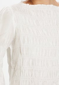 Blusa bianca e testurizzata con dettagli arricciati, maniche lunghe e scollatura rotonda, che crea una forma voluminosa. Tessuto leggero.