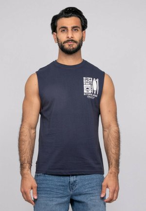 Homme aux cheveux noirs courts et barbe, portant un débardeur bleu marine avec un motif blanc et un jean bleu, debout devant un fond gris uni.