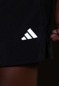 Falda deportiva negra con un logo de tres rayas blancas en el dobladillo, fabricada en una tela suave con un ligero brillo. Detalle de abertura lateral ajustable.