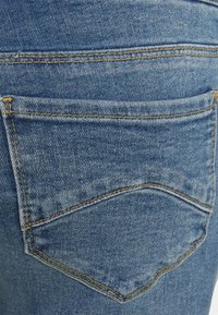 Os jeans de denim apresentam um bolso traseiro com costura amarela, tecido azul desbotado e um design de bolso curvado.