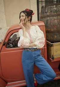 Witte blouse met rode borduursels en ballonmouwen, blauwe wijde jeans en een leopardprint riem. Rode schoenen en haar vastgebonden met een rode sjaal.