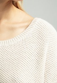Beige Strickpullover mit lockerer Maschenstruktur, weitem Ausschnitt und gerippten Bündchen, der ein strukturiertes Muster und einen subtilen Glanz zeigt.