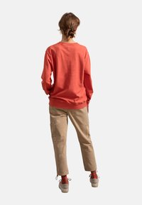 Felpa color corallo con polsini e fondo a coste, abbinata a pantaloni cargo beige dotati di tasche e segni di usura leggera. Calzature casual completano il look.