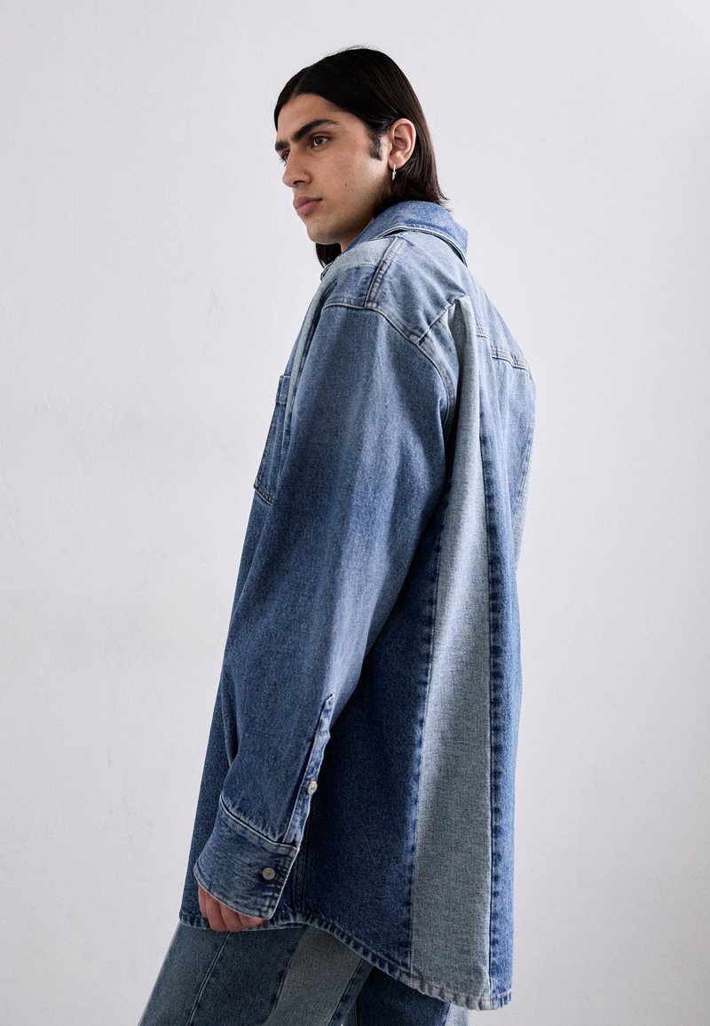 Camicia in denim con un design bicolore, con sezioni blu chiaro e scuro, chiusura con bottoni e maniche lunghe, dal taglio rilassato e dalla texture morbida.