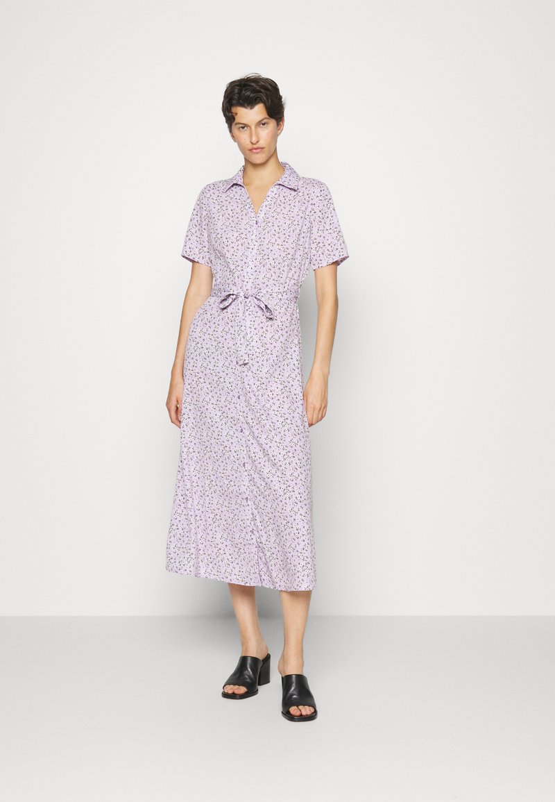 Vestido midi lilás claro com colarinho, mangas curtas, padrão floral, detalhe de cintura atada e fechamento frontal com botões. Combinado com mules pretos de ponta aberta.