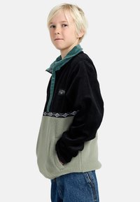 Fleece pulóver s čiernym zamatovým prešívaním, zeleným golierom a vzorovaným pásom. Obsahuje mäkkú sivú spodnú časť a prednú kapsu.