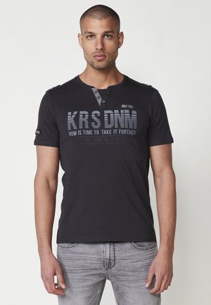 T-shirt nera in cotone con maniche corte, scollatura a girocollo, chiusura con due bottoni e stampa grafica sul davanti. Il testo presenta un mix di caratteri.