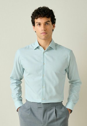 Man with curly hair wearing a light blue checkered button-up shirt and gray trousers, standing with hands in pockets against plain background.