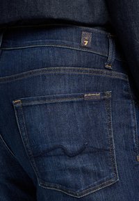 Jeans in denim blu scuro con tasca posteriore decorata da dettagli di cucitura, un piccolo etichetta di marca e una texture liscia.