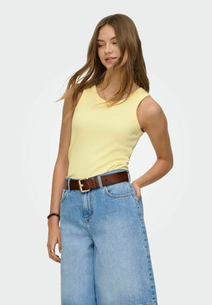 Donna che indossa un top giallo senza maniche infilato in jeans azzurri a vita alta con cintura marrone, che guarda di lato con una mano nella tasca posteriore.