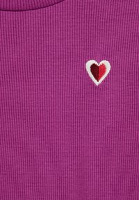 Top en tissu ribbé violet avec un petit design de cœur brodé en rouge et blanc sur la poitrine, avec une texture prononcée visible.