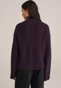 Femme aux longs cheveux noirs portant un pull côtelé violet foncé et un pantalon noir, se tenant de dos à la caméra contre un fond uni.