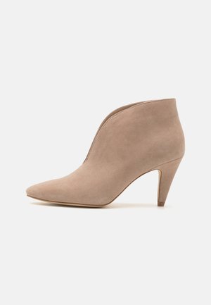 Beige ruskind dameankelstøvle med spids tå og mellemstor kegleformet hæl på ensfarvet hvid baggrund.