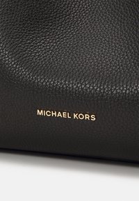 Černý strukturovaný kožený povrch s vyraženým zlatým logem "MICHAEL KORS" blízko spodního okraje kabelky nebo doplňku.