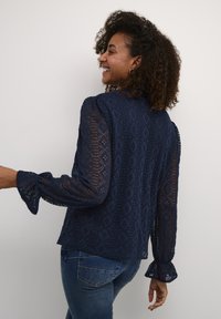 Blusa in pizzo blu navy con maniche lunghe, un motivo a diamante testurizzato, polsini a scallop e orlo arrotondato. Indossata con jeans in denim blu.