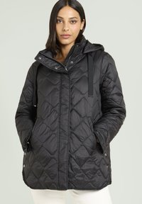 Donna che indossa una giacca a cappuccio trapuntata nera con bottoni a pressione e zip, con le mani nelle tasche, in piedi di fronte a uno sfondo chiaro e semplice.