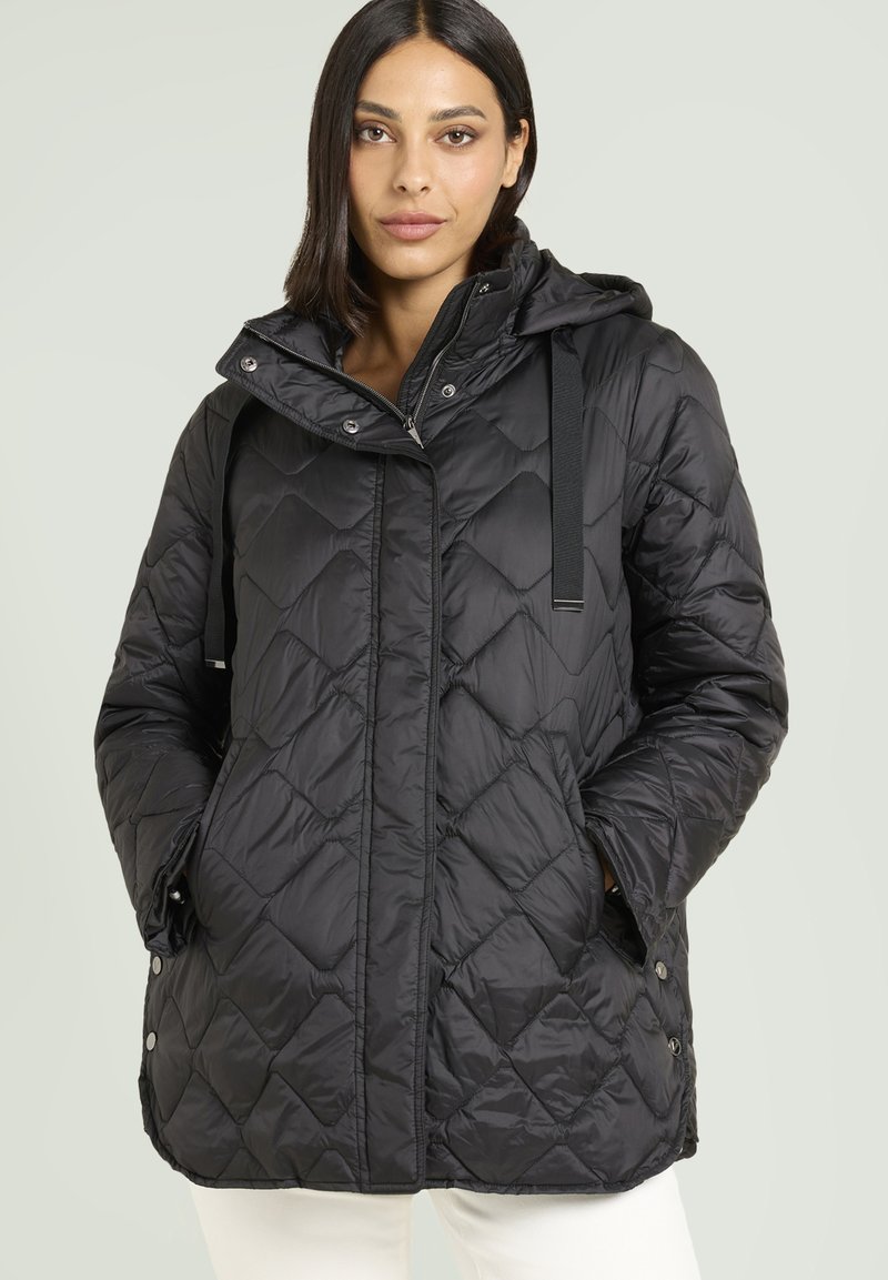 Donna che indossa una giacca a cappuccio trapuntata nera con bottoni a pressione e zip, con le mani nelle tasche, in piedi di fronte a uno sfondo chiaro e semplice.