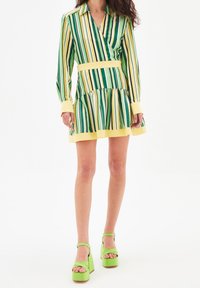 Vestido cruzado de manga larga en tela de rayas verdes y amarillas con falda con volantes. Acompañado de zapatos de plataforma verdes con tiras en el tobillo.