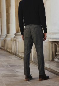 Gray tweed trousers with a tailored fit, featuring two back pockets, paired with brown, lace-up shoes, and a black long-sleeve top.